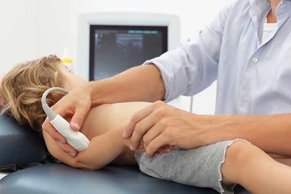 Linear ultrasound probe placed over the lateral elbow of a clothed toddler, pointing toward the hand, to obtain the radiohumeral view.