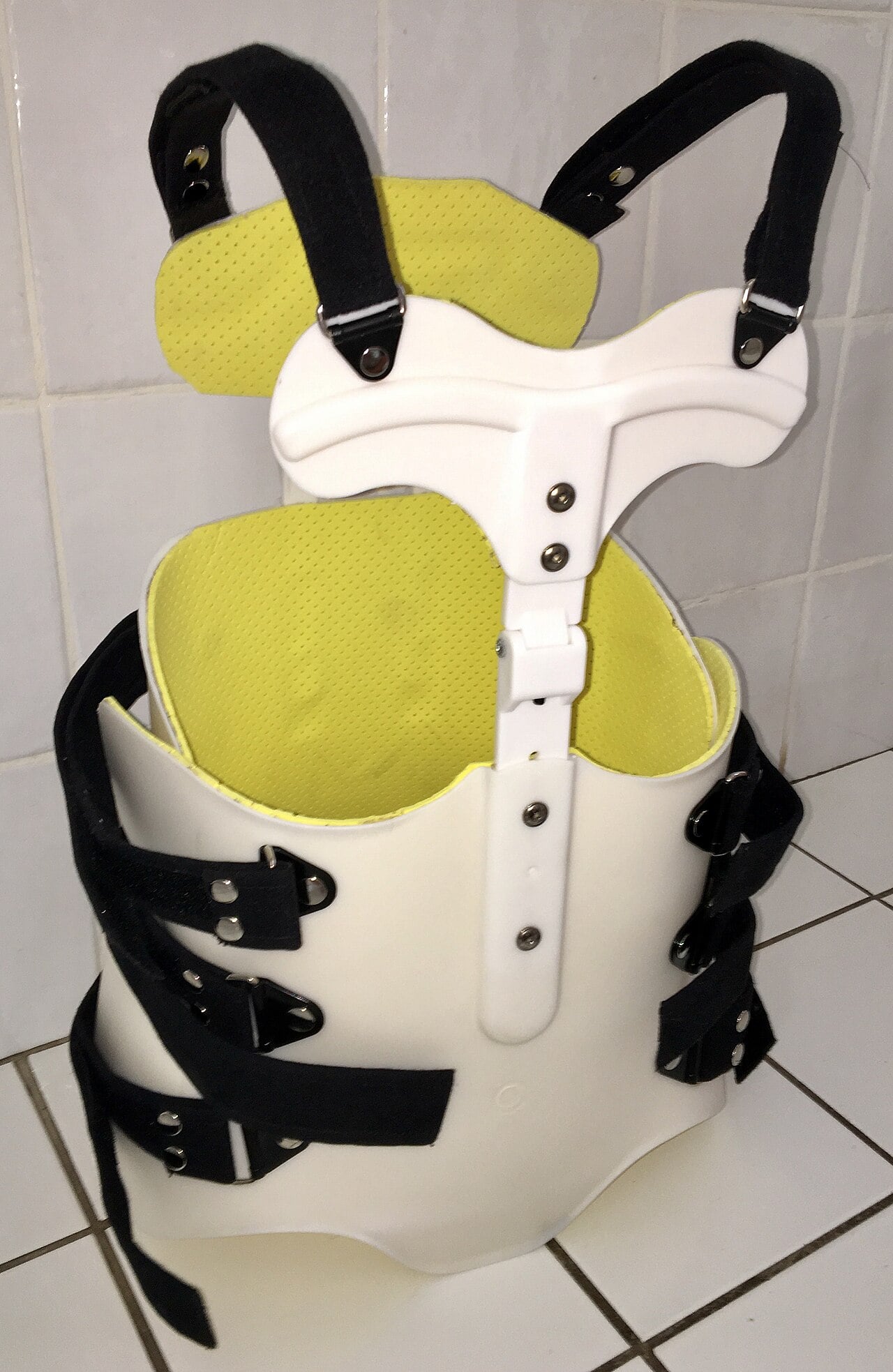 Thoracolumbosacral orthosis standing empty, with a foam-lined plastic shell, side straps and a T-shaped upper plate on shoulder straps.