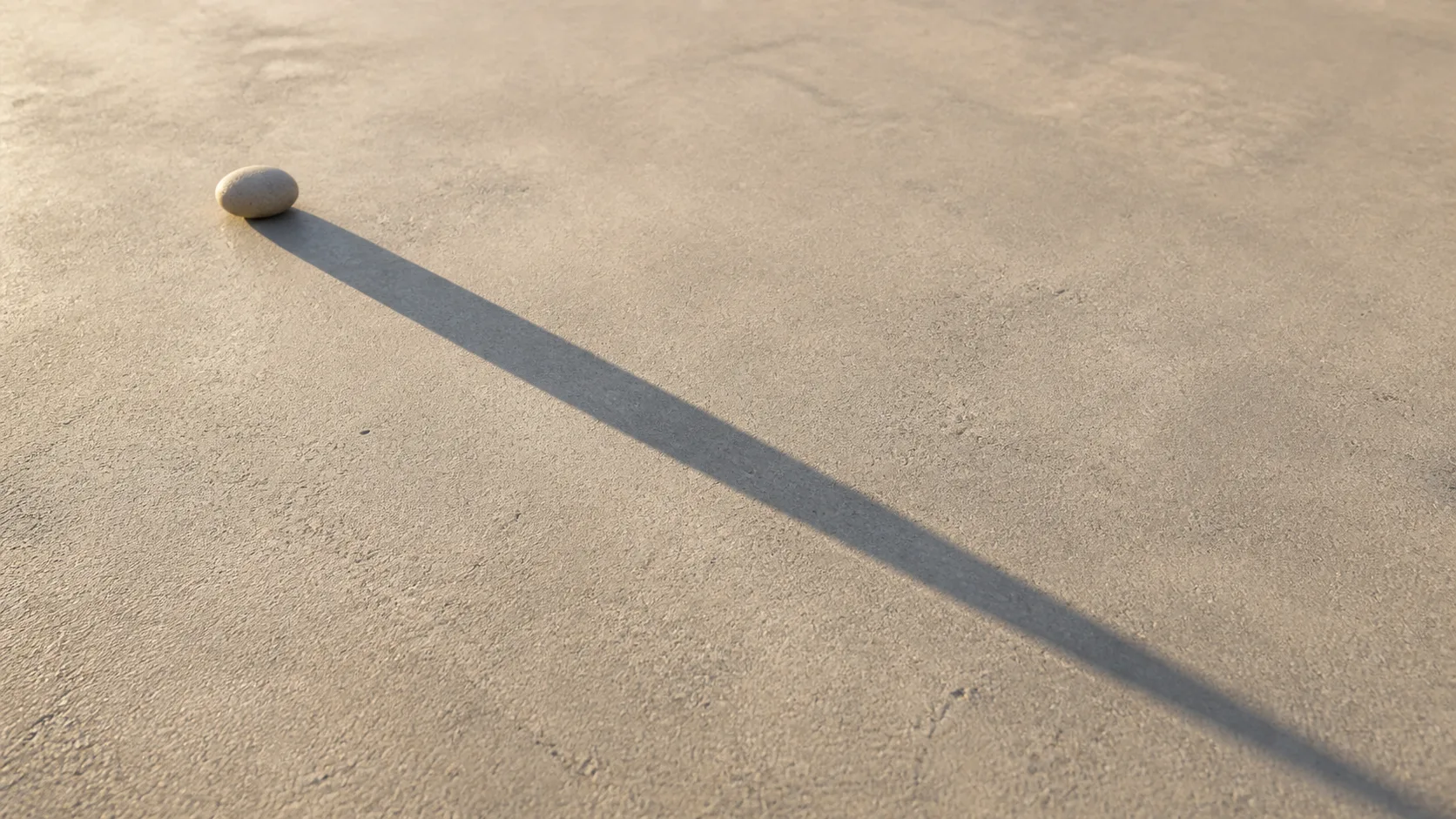 A single small object casting a very long shadow across a pale concrete floor in low raking sunlight