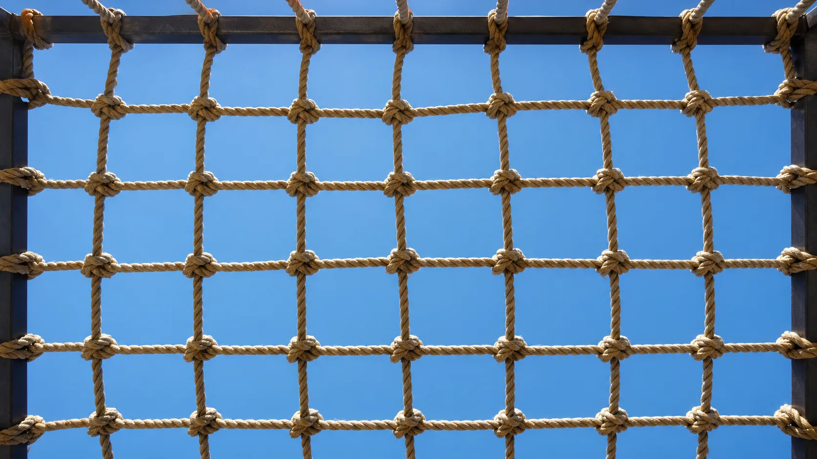 A taut knotted rope safety net stretched across a frame photographed from below against an open bright sky