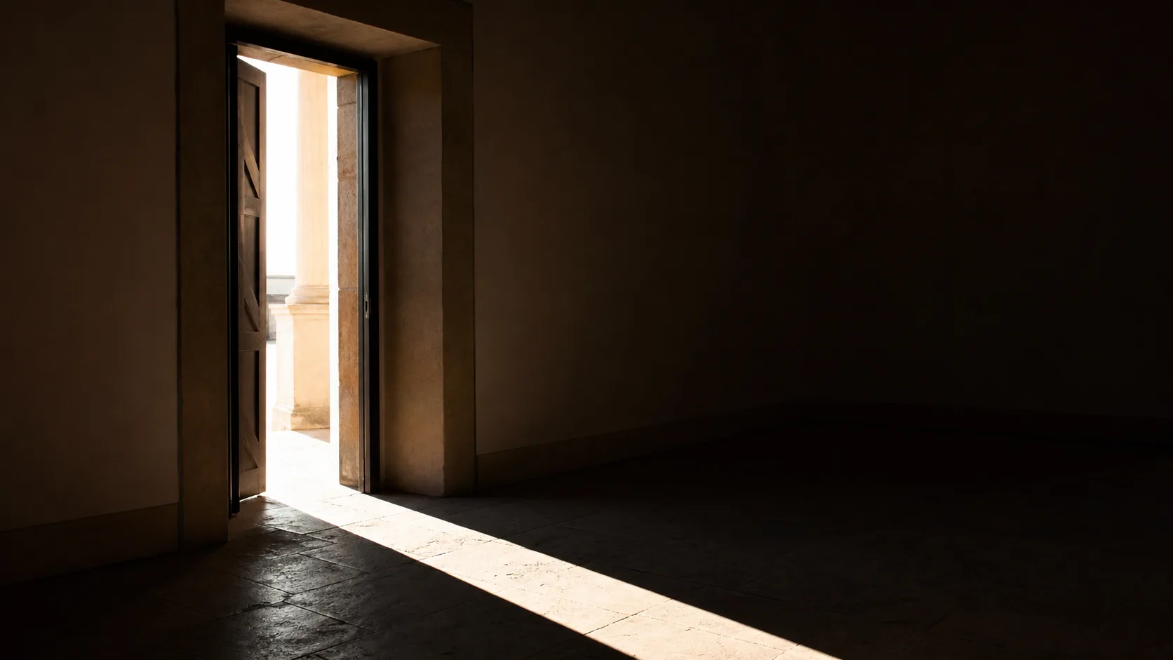 An open door with bright daylight spilling across a dark floor into a shadowed room