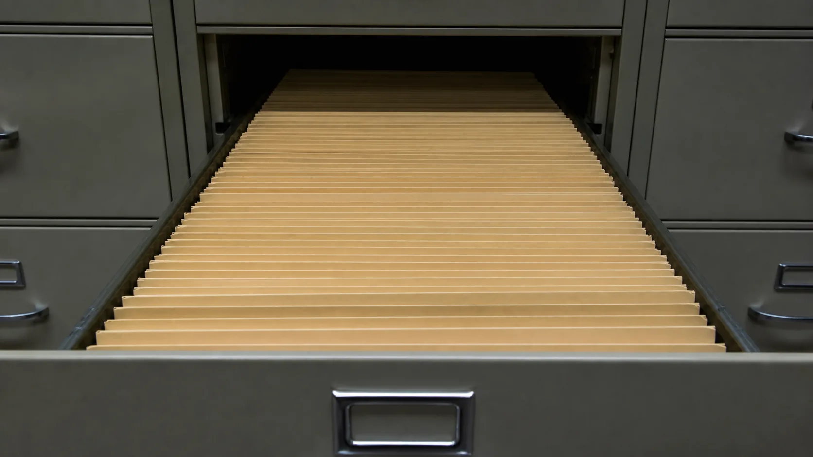 An open archive drawer packed with dozens of identical buff folders receding into shadow