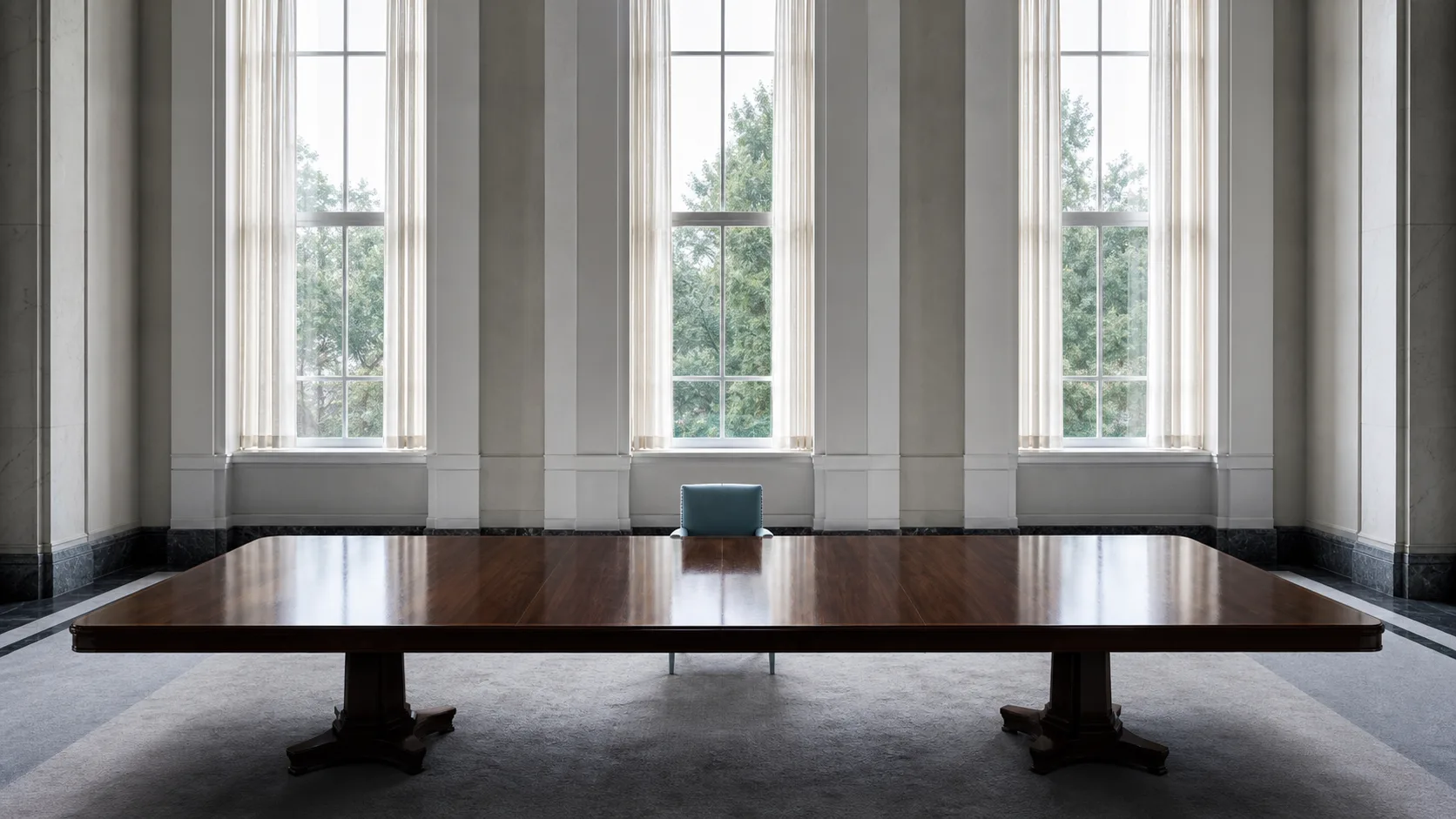 A single upholstered chair at the centre of one side of a long polished table in an empty formal room